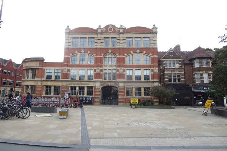 A historic red brick building with large windows, flanked by other buildings and bicycles parked in front. The area is paved and includes signage.